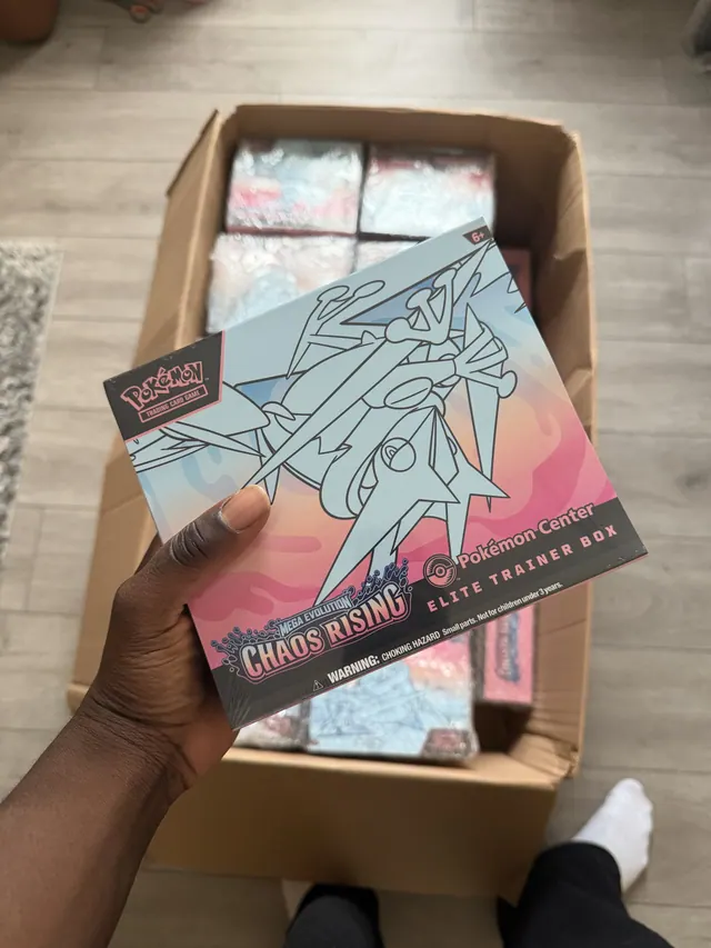 A hand holding a colorful Pokémon Elite Trainer Box above an open delivery carton