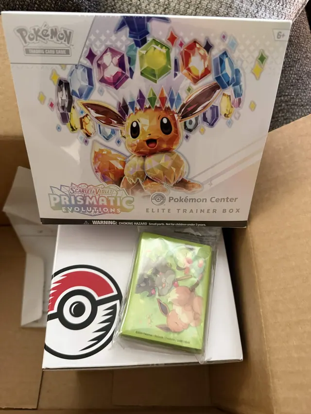 An Eevee-themed Prismatic Evolutions box above an open carton with card accessories