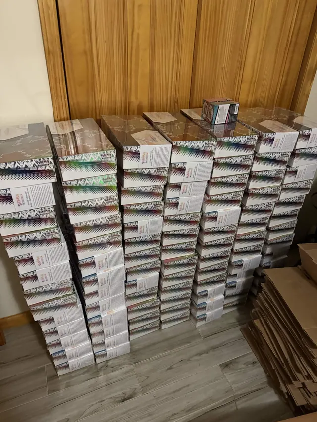 Tall stacks of sealed Pokémon card boxes arranged beside a wooden wardrobe.
